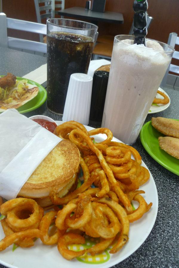 A juicy burger with crisp bacon and a side of seasoned curly potato fries sits in front of a barbeque chicken wrap and a plate of apple turnovers with a soda and chocolate milkshake to wash it all down.