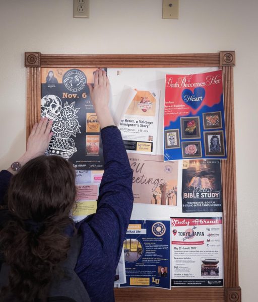 A college student reaching up to tape a poster to the top left corner of a full bulletin board.