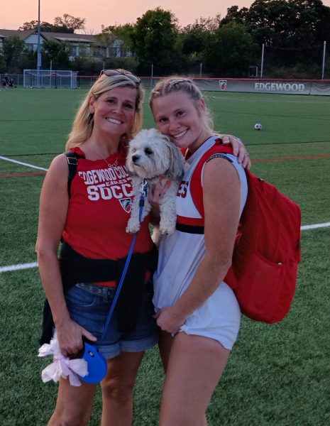 Two smiling woman standing together. The woman on the left wears a red shirt that reads "Edgewood Soccer," and the woman on the right holds a small white dog.