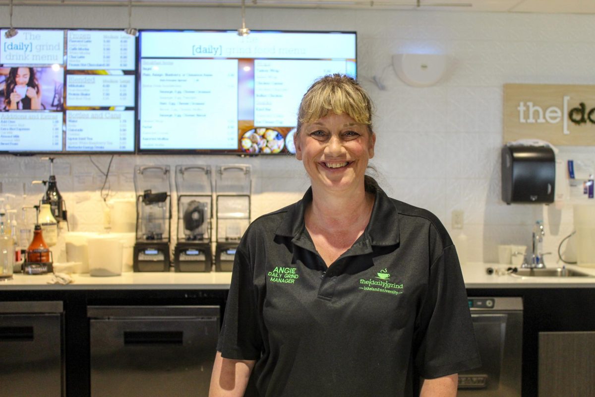 A smiling woman stands in front of drink blenders and two TV screens with drink and food menu items.