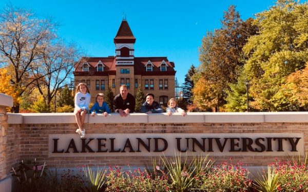 A family of five sitting on a brick wall with large letters that read "Lakeland University." In the background is Lakeland's Old Main.